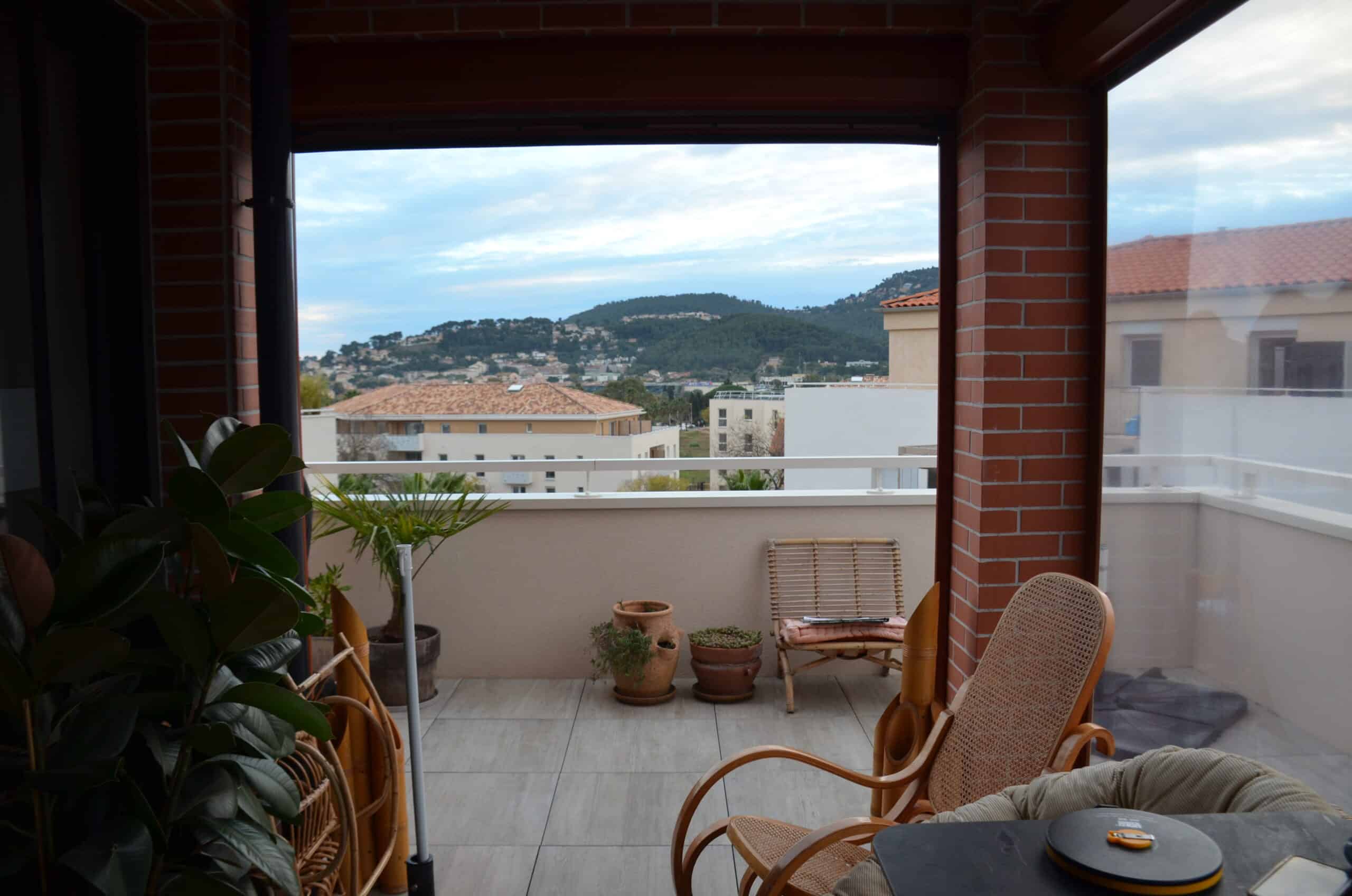 Balcon avec mobilier en rotin, plantes en pot et vue panoramique sur les collines et toits de la ville.
