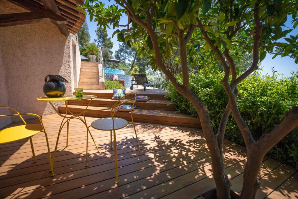 Terrasse bois et mobilier jaune : Détente au jardin avec piscine. Terrasse en bois ensoleillée et jardin luxuriant. Mobilier extérieur jaune vif sous un arbre, menant aux escaliers d'une piscine en mosaïque bleue.