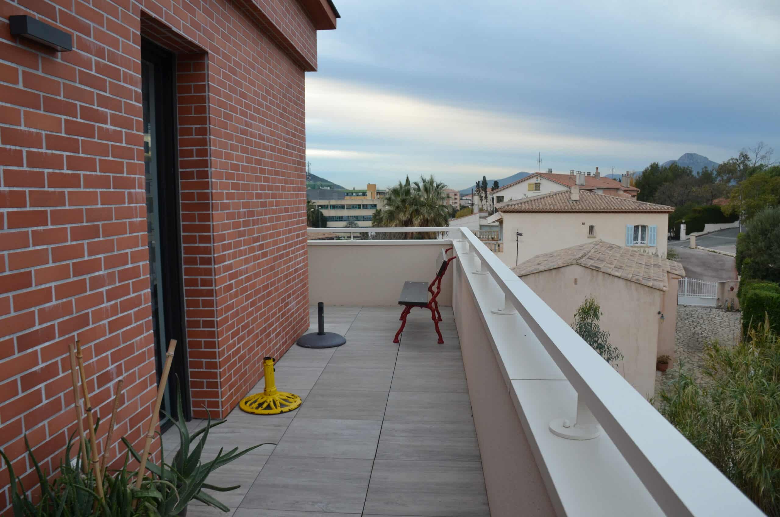 Terrasse moderne avec mur en briques rouges, banc rouge et vue panoramique sur les toits et montagnes environnantes.