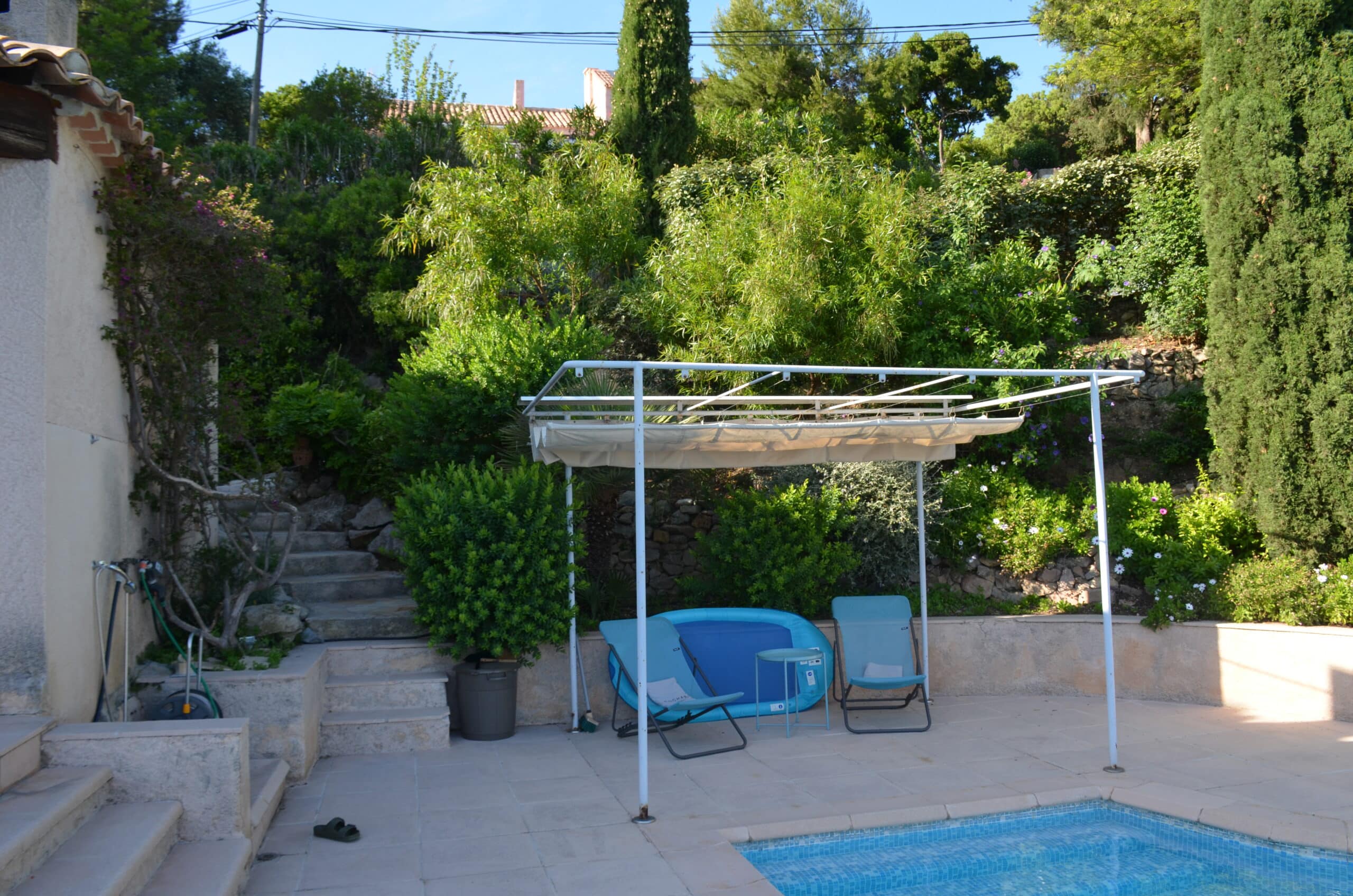 Terrasse de piscine ensoleillée avec pergola, transats bleus et jardin luxuriant méditerranéen avec escalier en pierre.