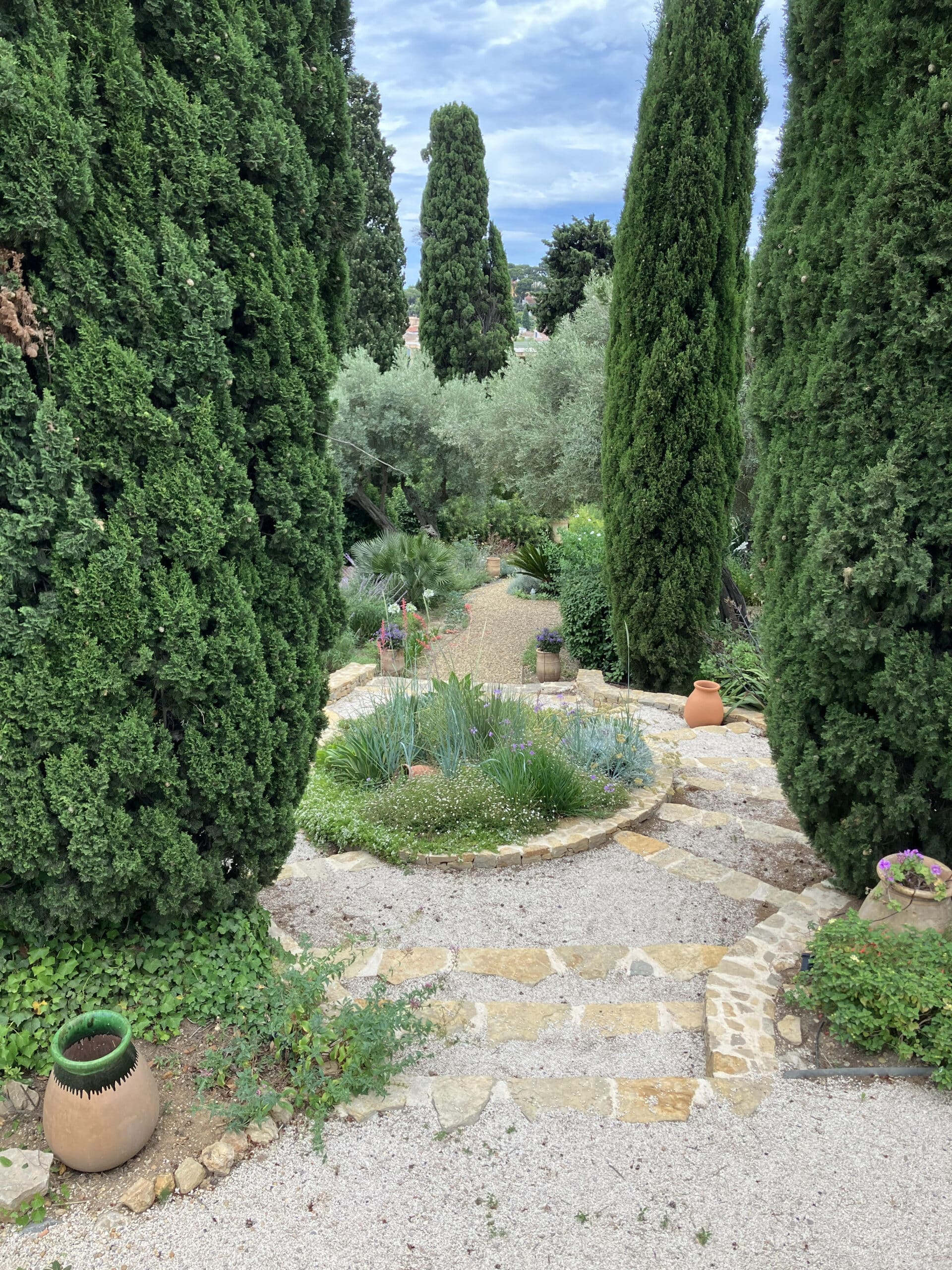Jardin méditerranéen avec cyprès élancés, escalier en pierre et pots en terre cuite sur un sentier de gravier.