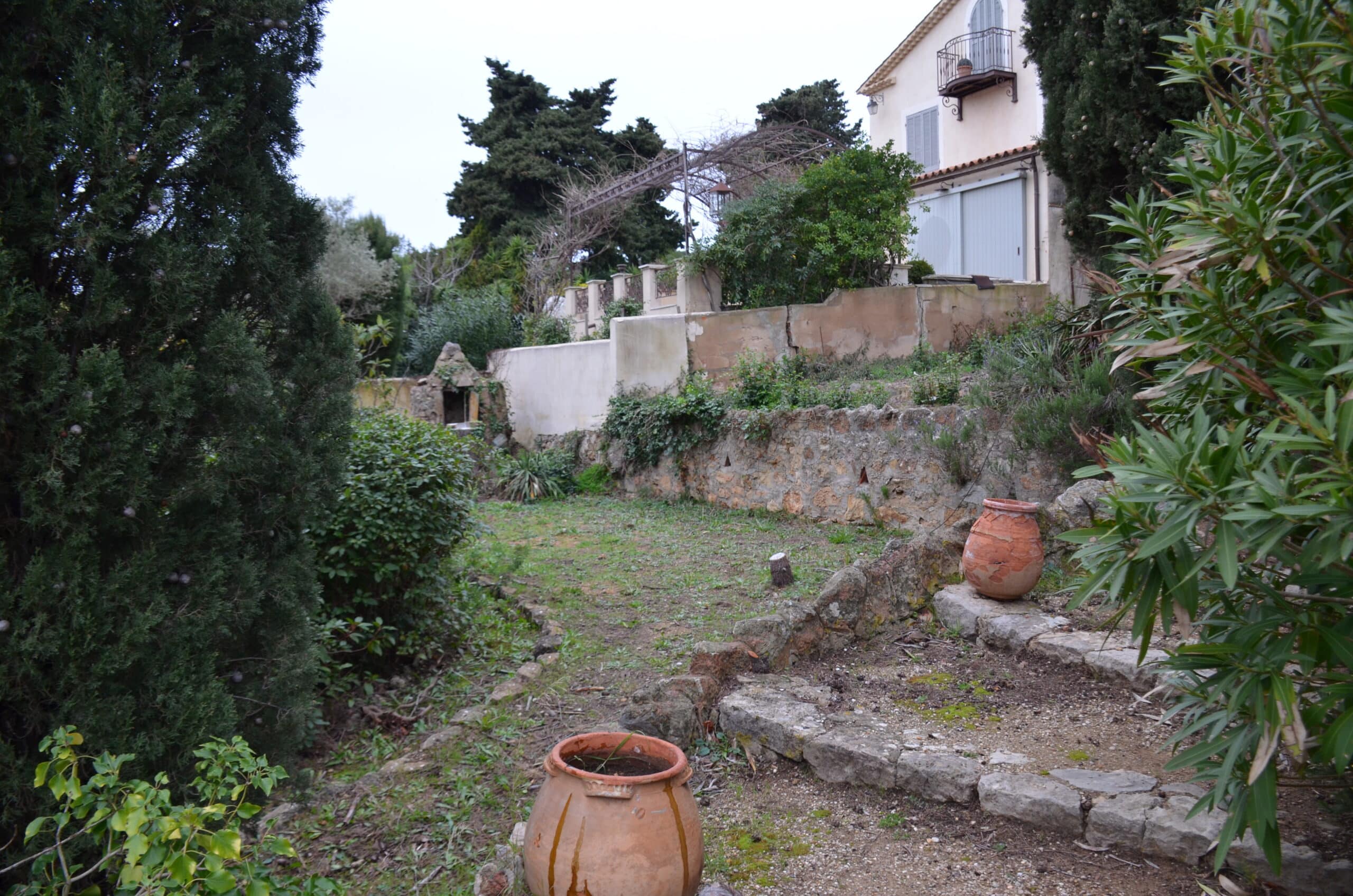 Jardin en terrasses avec jarres en terre cuite et murets en pierre devant une villa méditerranéenne.