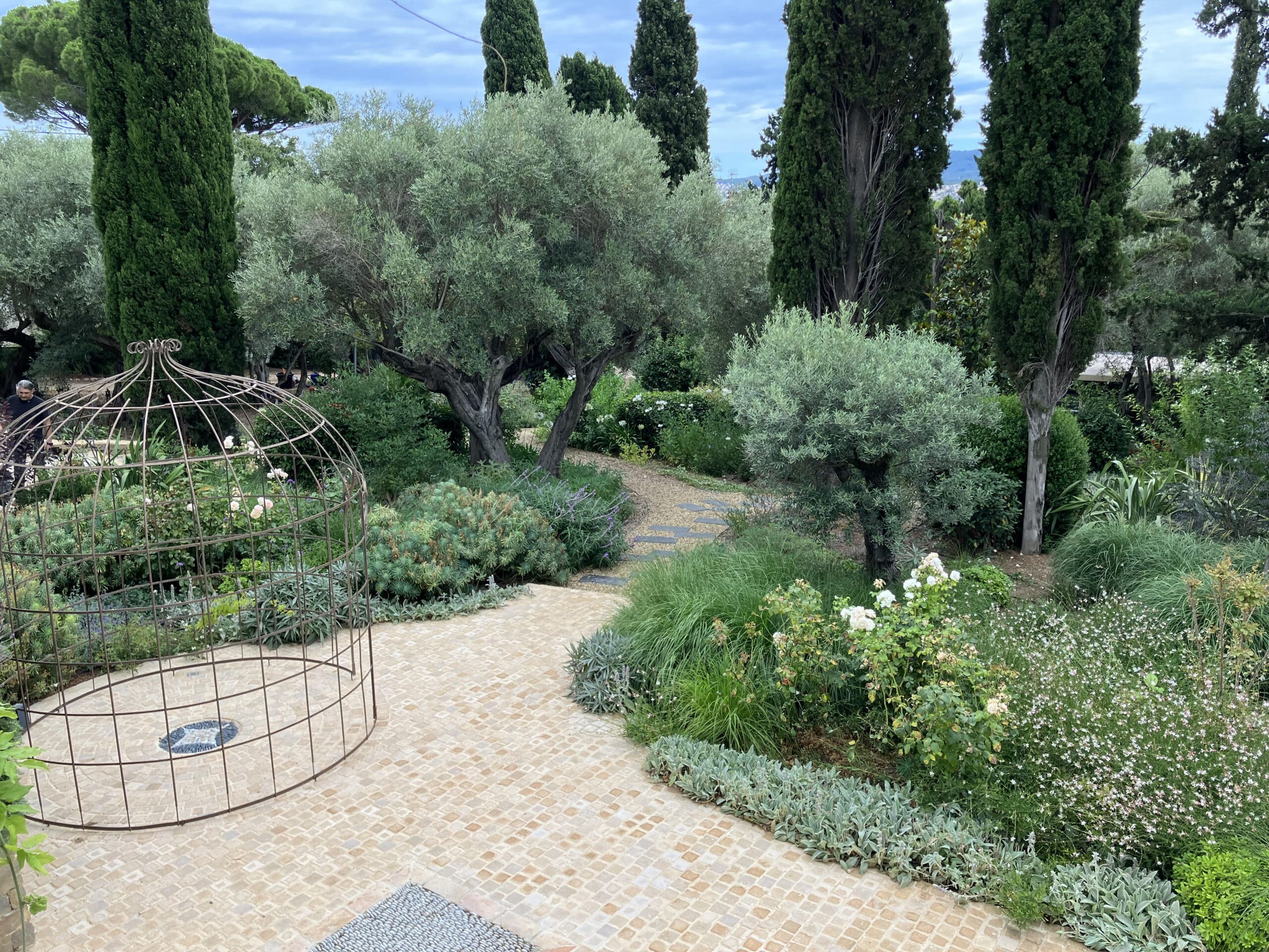 Jardin méditerranéen avec structure en fer forgé, oliviers et cyprès bordant une allée pavée en pierre.