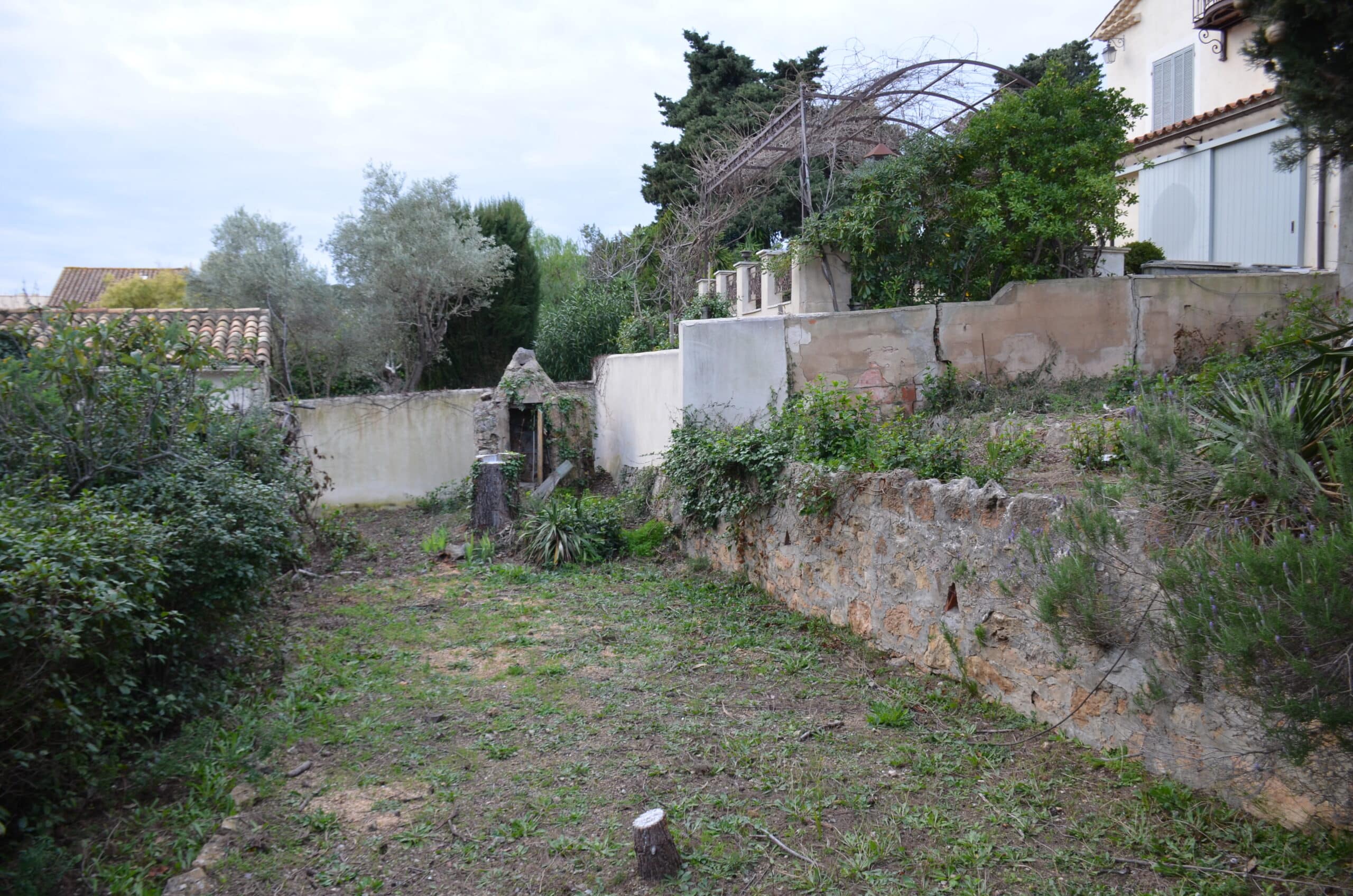 Jardin en terrasses avec muret en pierre, végétation sauvage et petite cabane en pierre sous un ciel nuageux.