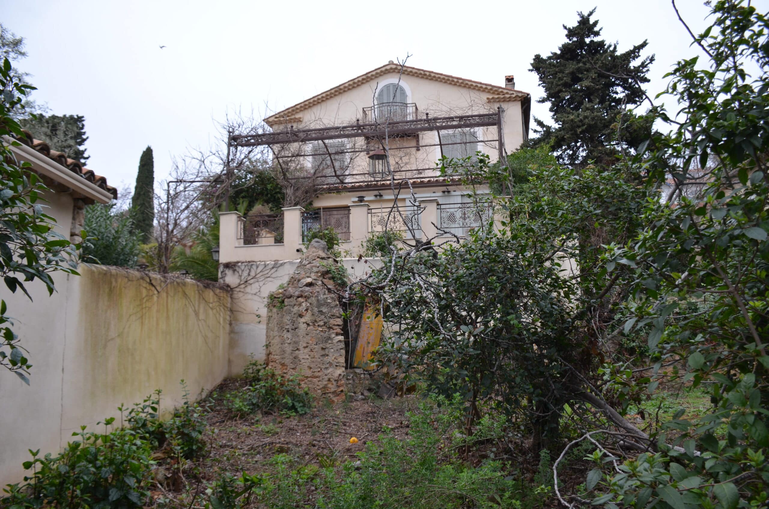 Maison à étage avec balcon en fer forgé, jardin sauvage et mur en pierre ancienne sous un ciel couvert.