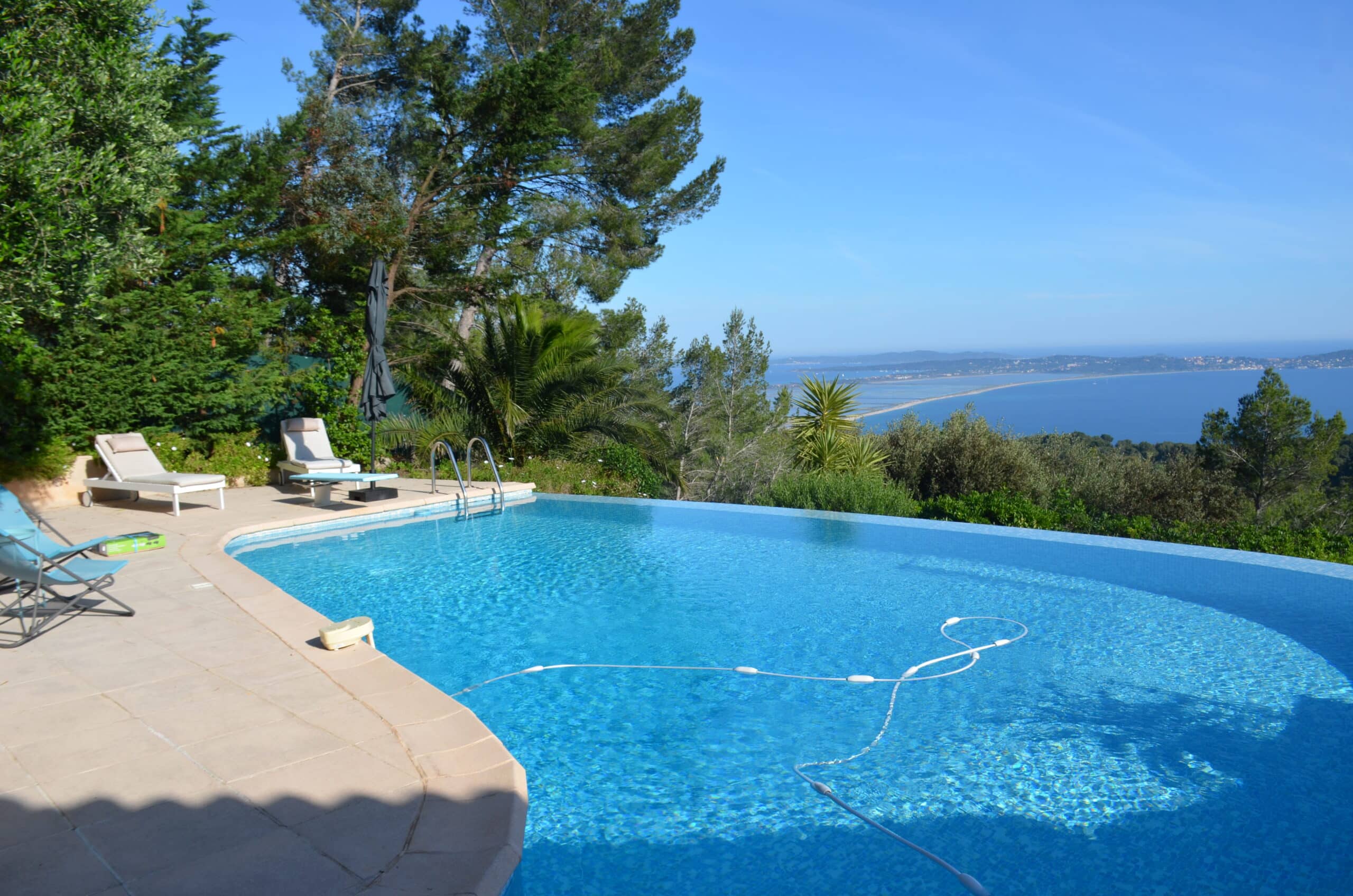 Piscine à débordement avec vue panoramique sur mer, transats et jardin méditerranéen sous un ciel bleu.