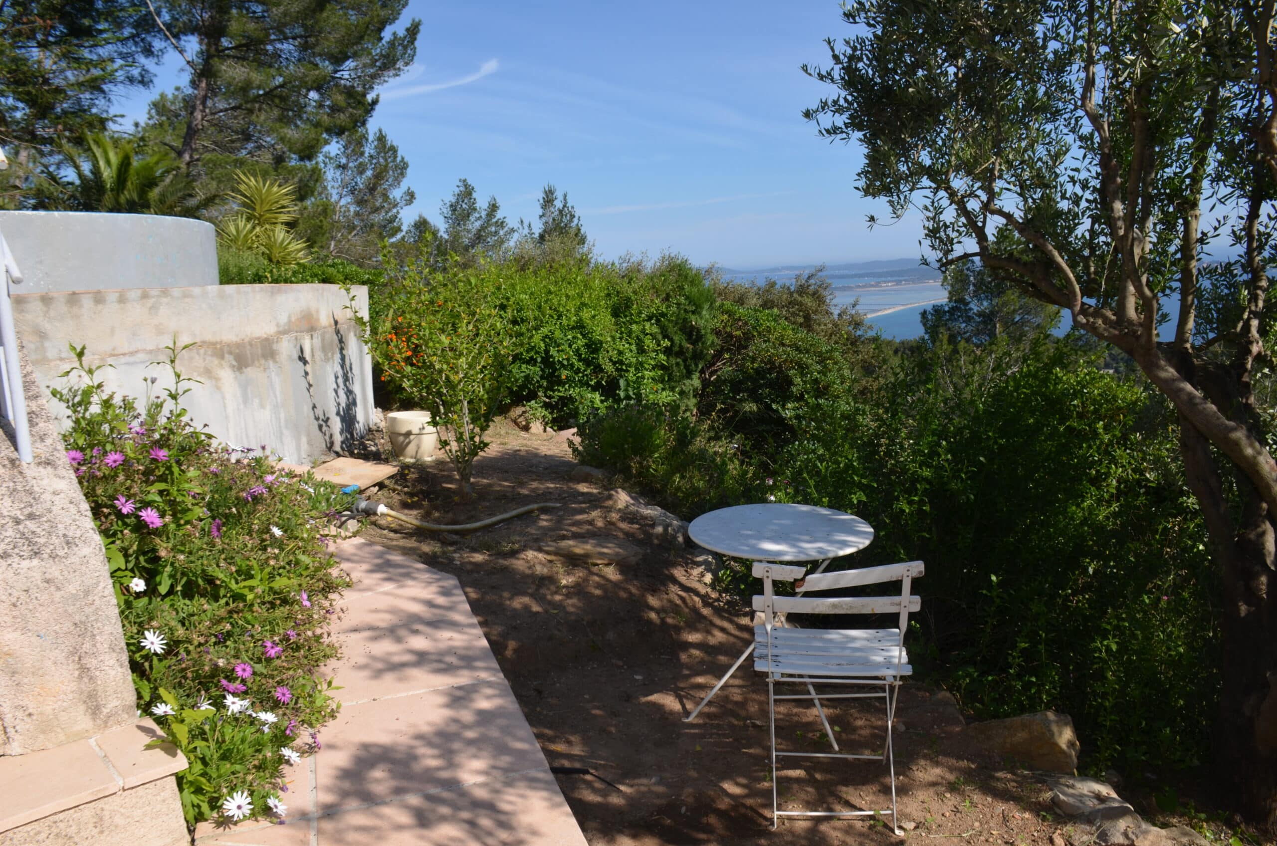 Terrasse ensoleillée avec table blanche, fleurs violettes et vue panoramique sur la mer Méditerranée.