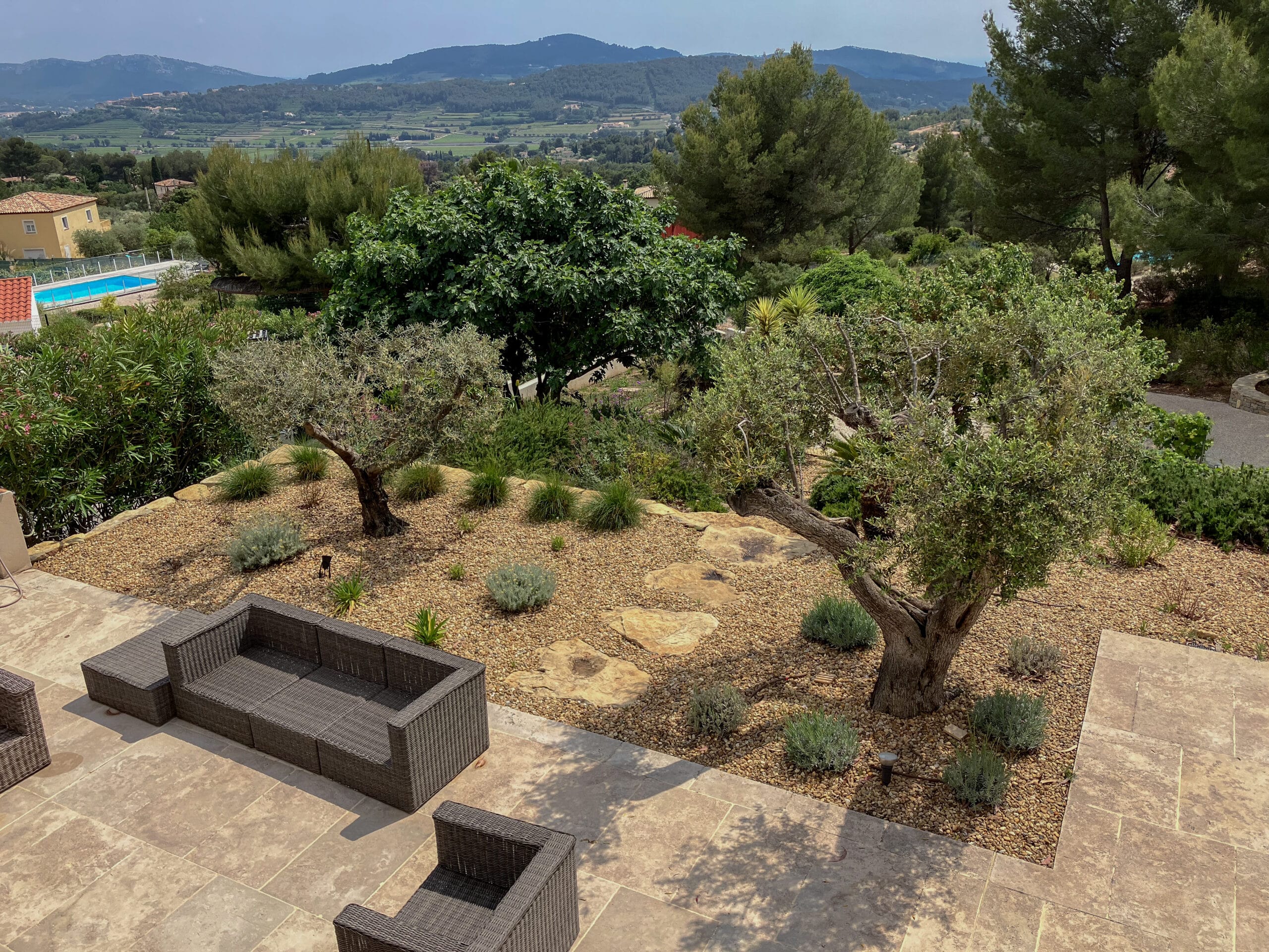 Terrasse en pierre avec canapé en résine, oliviers, jardin paysager et vue panoramique sur les montagnes méditerranéennes.