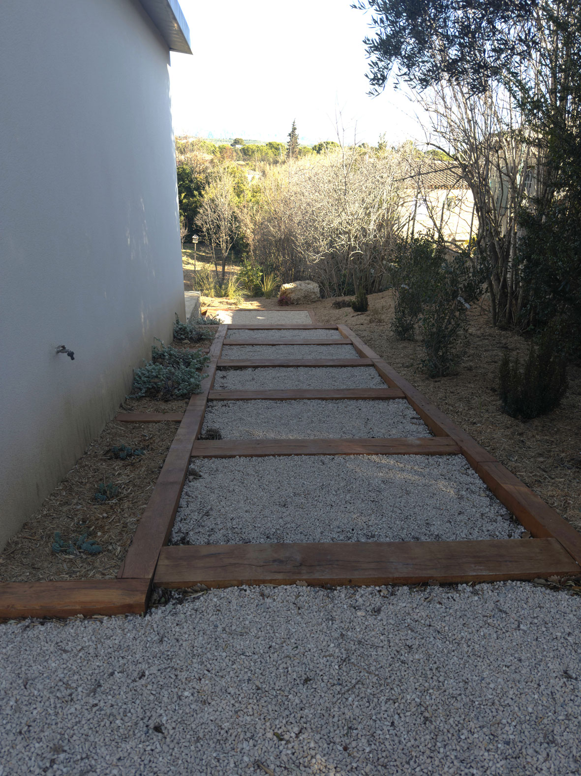 Allée de jardin paysagée avec traverses de bois et gravier blanc longeant une façade de maison moderne.