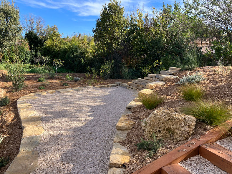 Jardin paysager avec chemin en gravier clair, bordures en pierre naturelle, paillage en bois et escalier de jardin.