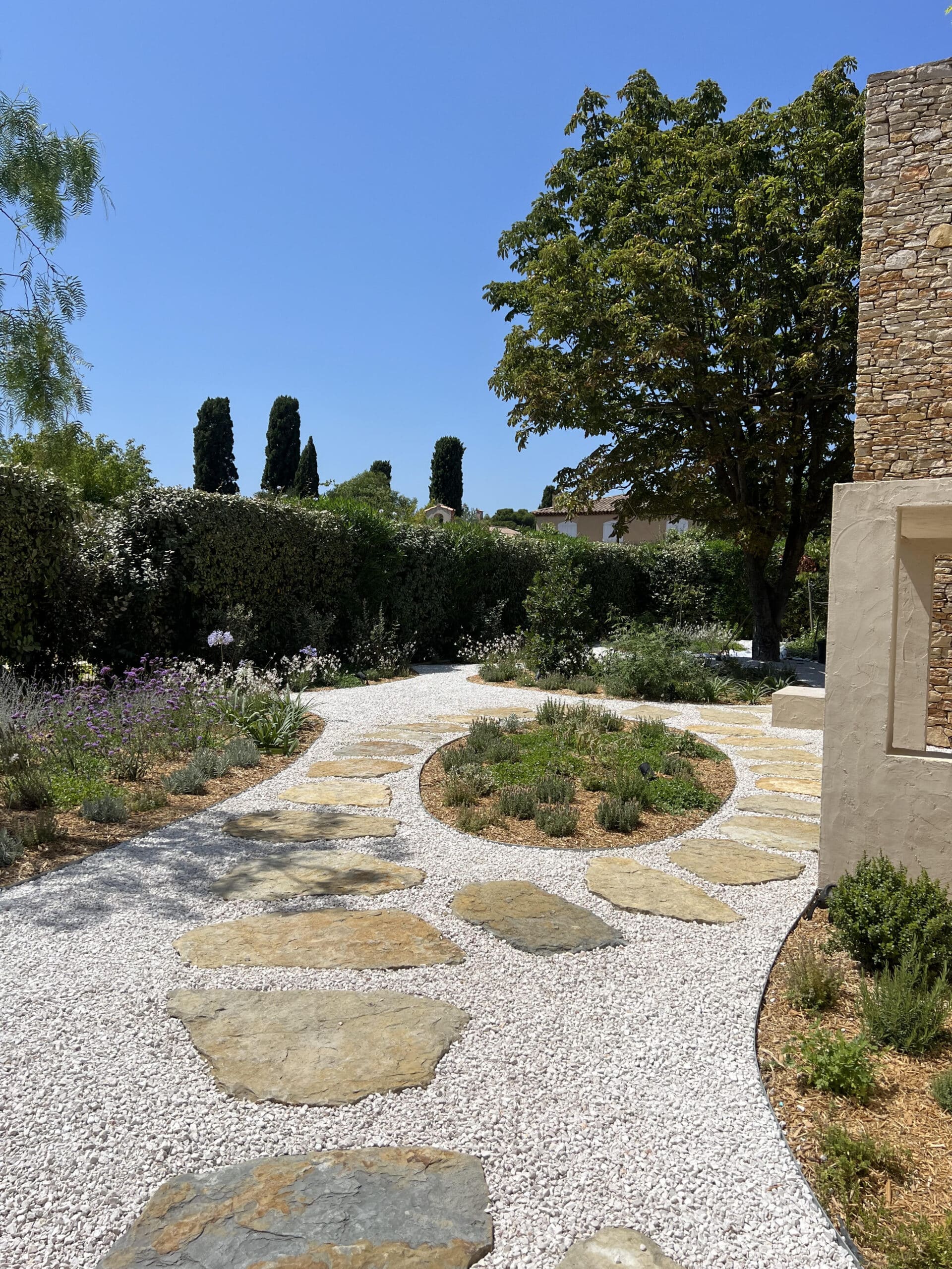 Jardin paysager avec allée en gravier blanc, dalles de pierre naturelles et massif circulaire fleuri.