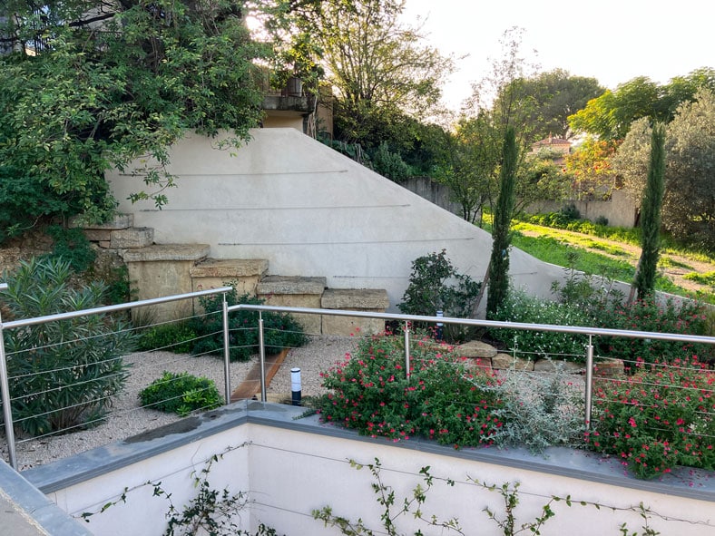 Jardin moderne avec escalier en pierre, muret blanc rainuré, garde-corps inox et fleurs rouges paysagères.