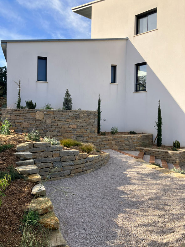 Jardin moderne avec murs en pierre sèche, allée en gravier clair et maison contemporaine sous le soleil.