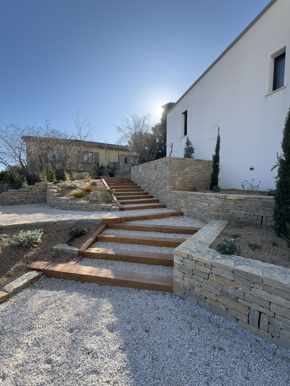 Escalier de jardin en bois et gravier bordé de murets en pierre sèche devant une maison blanche moderne.