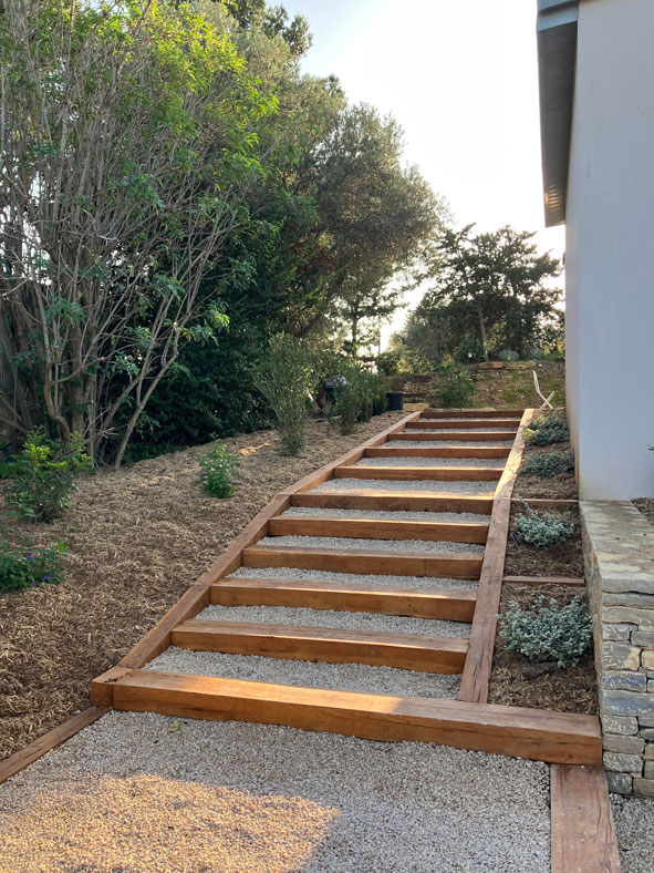 Escalier de jardin en traverses de bois et gravier blanc longeant une pente avec aménagement paysager naturel.