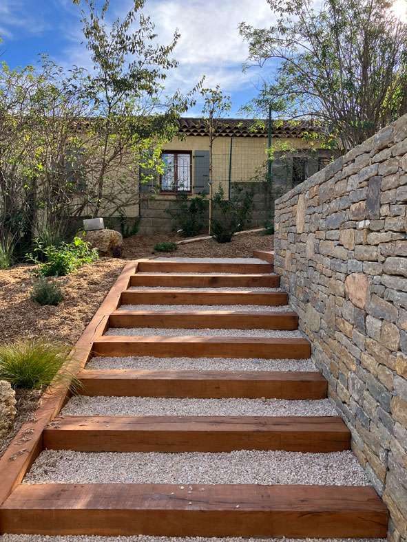 Escalier extérieur en poutres de bois et gravier blanc longeant un mur en pierre sèche dans un jardin méditerranéen.