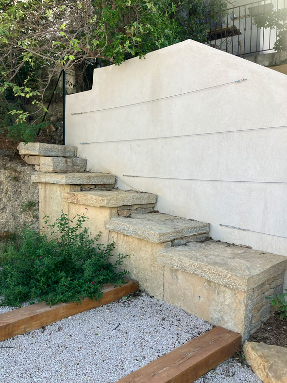 Escalier en pierre naturelle et mur blanc avec fils de palissage pour plantes grimpantes dans un jardin paysagé.