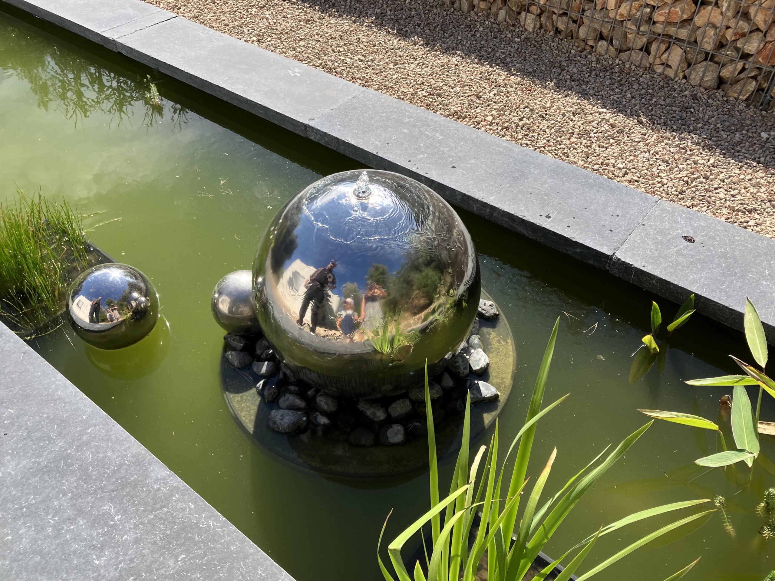 Fontaine sphérique en inox avec reflets de visiteurs dans un bassin de jardin verdoyant.