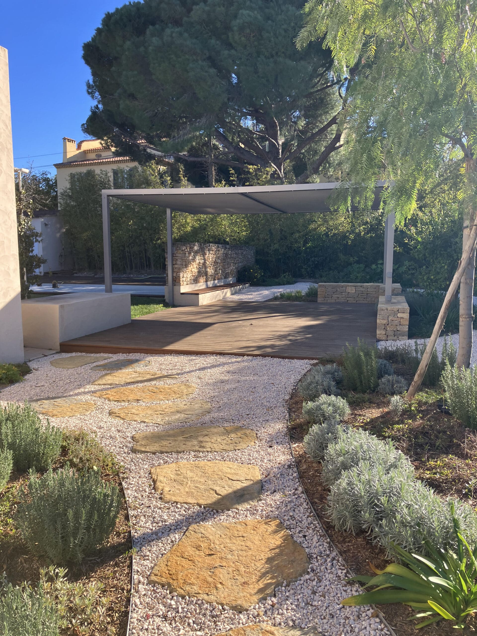 Jardin méditerranéen avec pergola, terrasse en bois, pas japonais en pierre et chemin en gravier blanc ensoleillé.