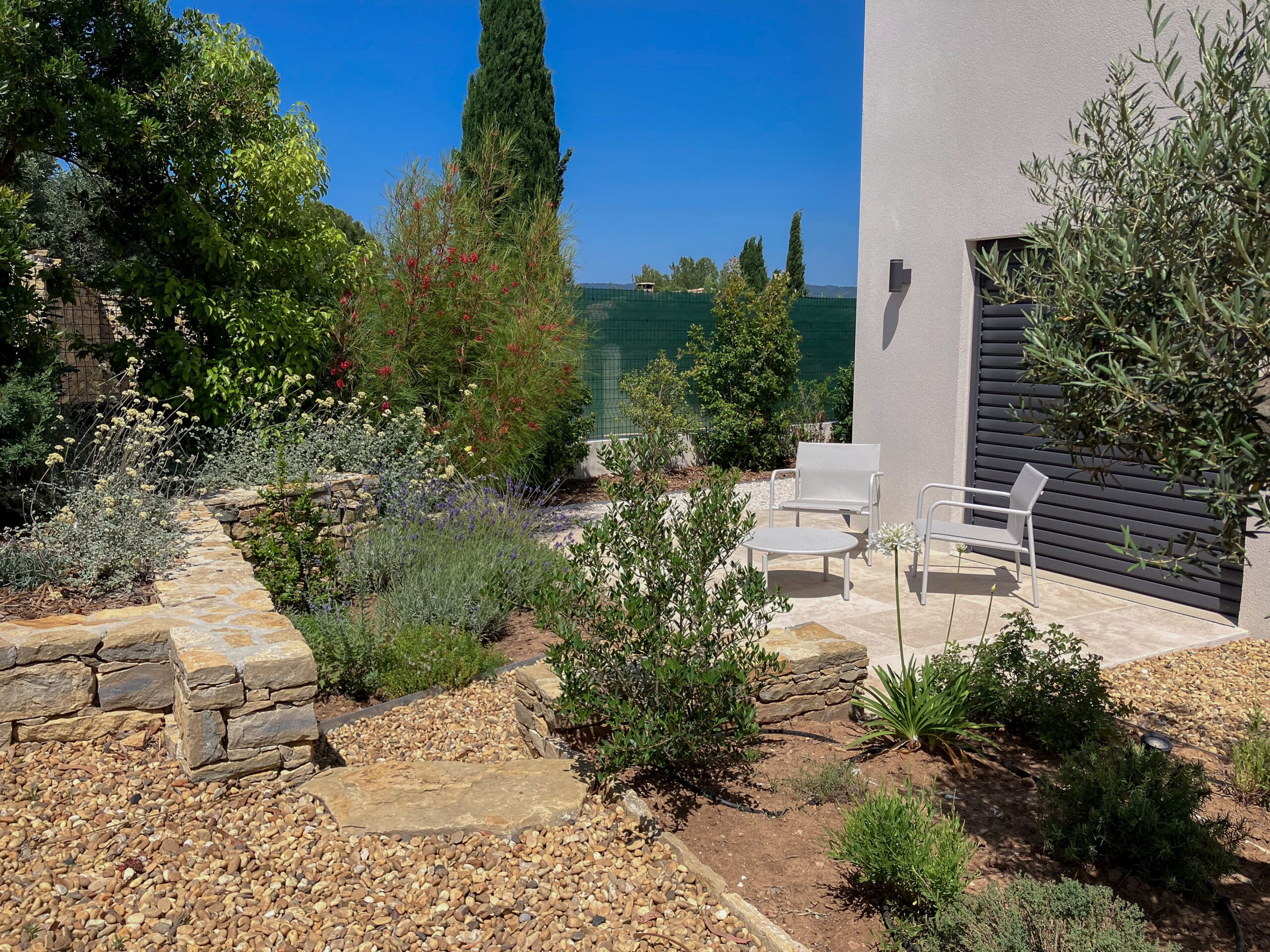 Jardin moderne avec terrasse en pierre, fauteuils gris, muret de pierre sèche et végétation méditerranéenne.