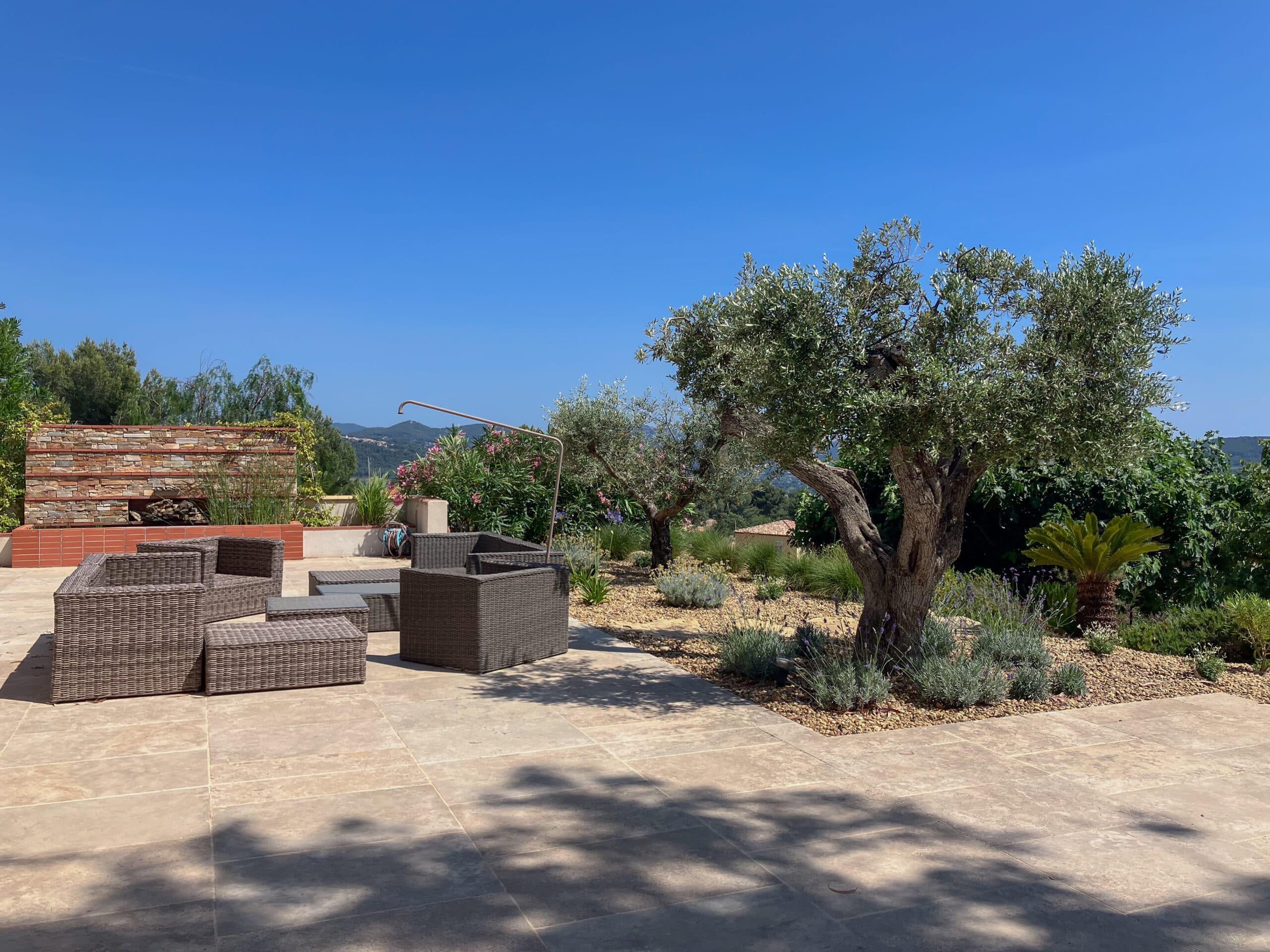Terrasse avec salon en résine tressée, grand olivier et vue sur les montagnes sous un ciel bleu dégagé.