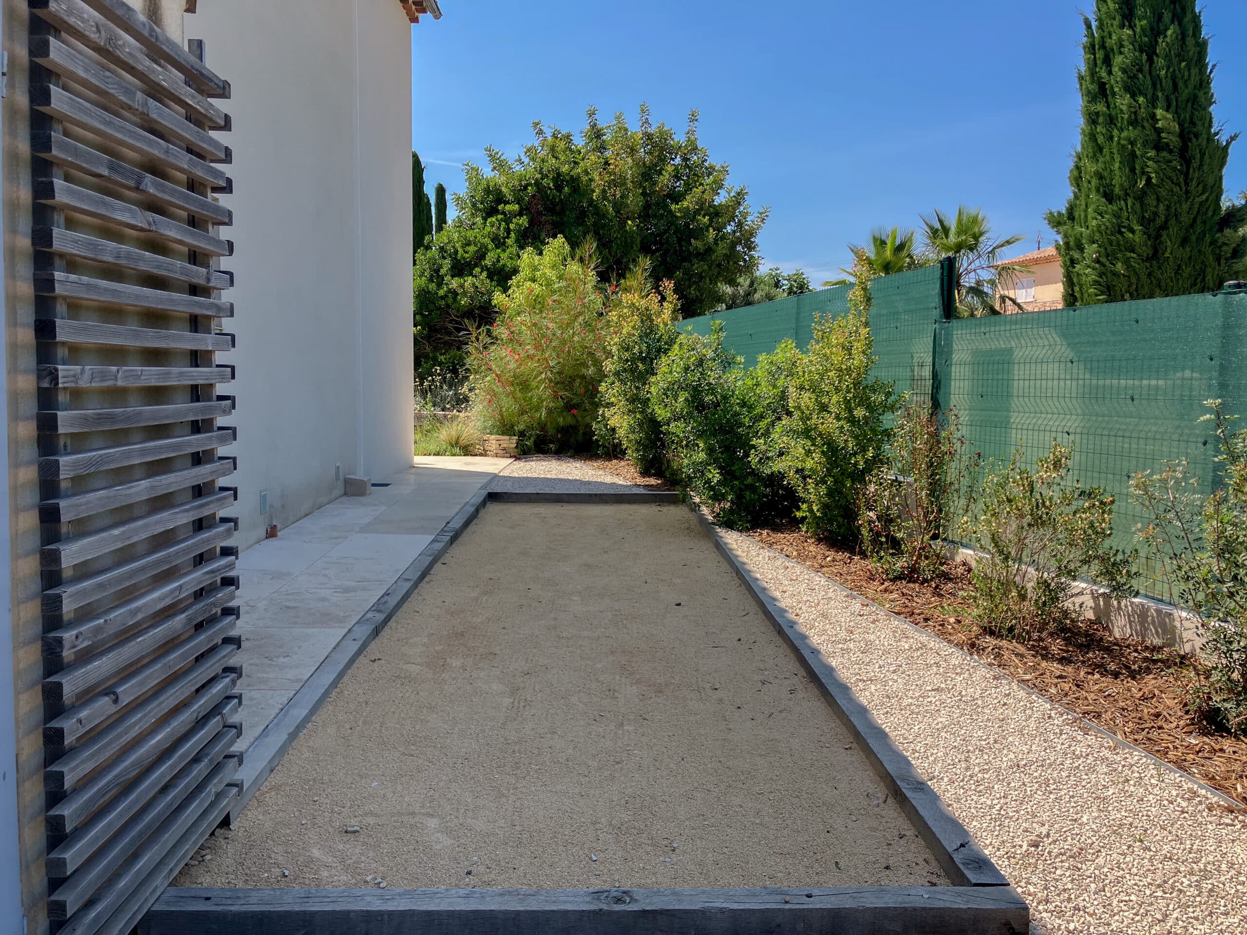Terrain de pétanque en sable bordé de bois dans un jardin ensoleillé à côté d'une maison moderne.
