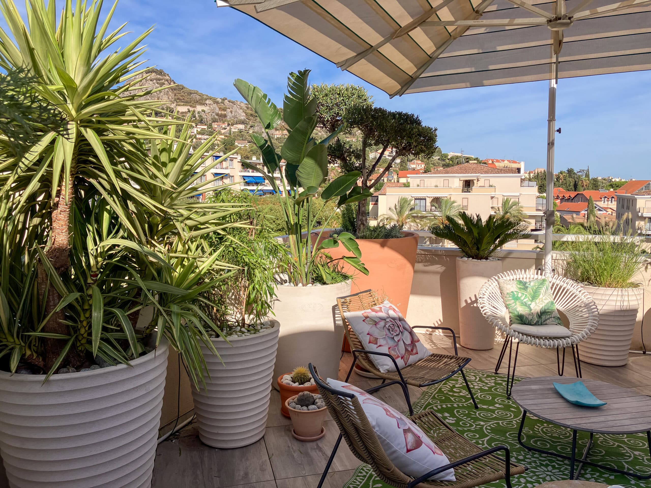 Terrasse fleurie avec chaises en osier, plantes tropicales et vue panoramique sur une montagne et la ville.
