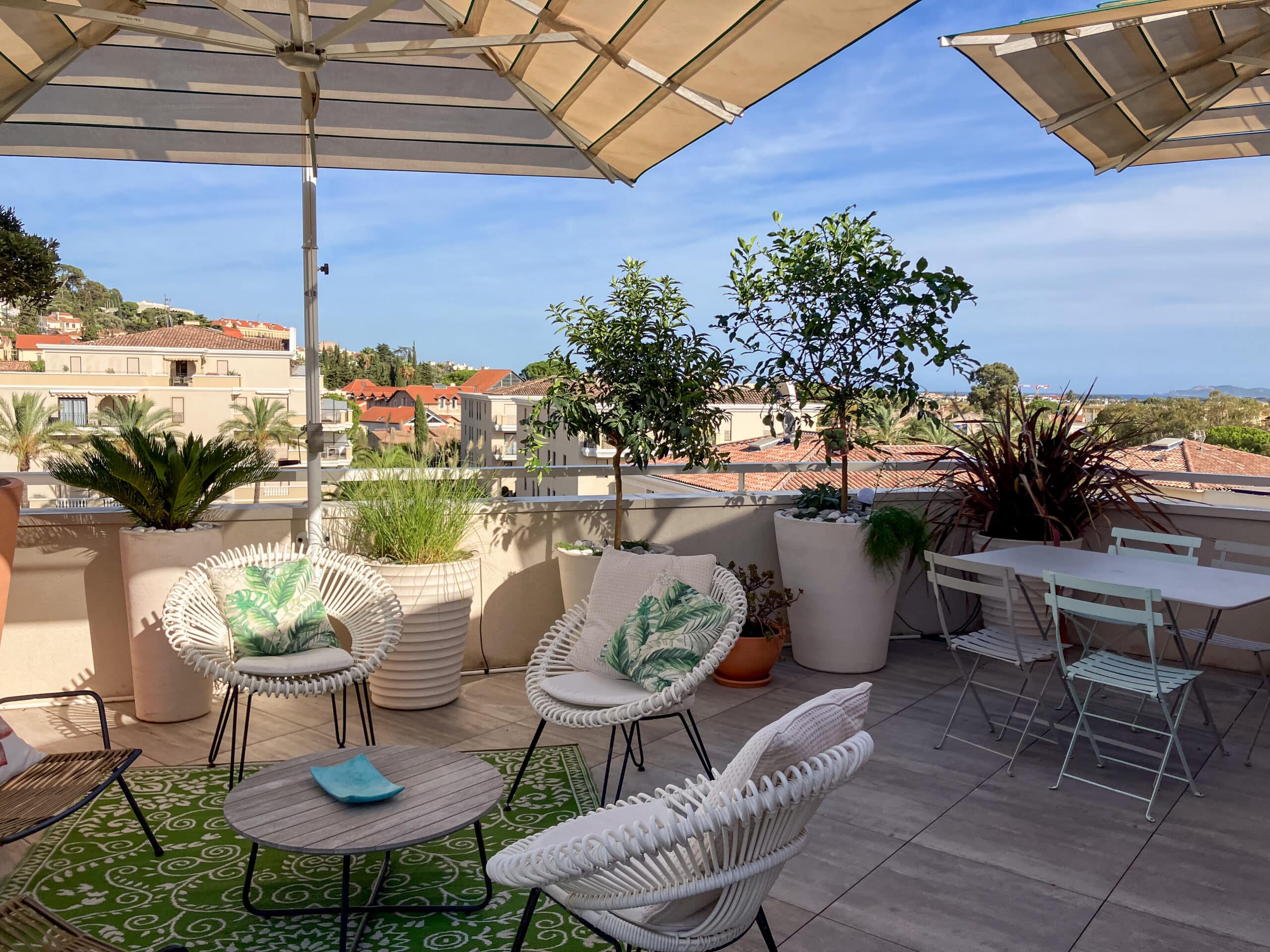 Terrasse ensoleillée avec chaises en rotin blanc, plantes vertes et vue panoramique urbaine sous un grand parasol.