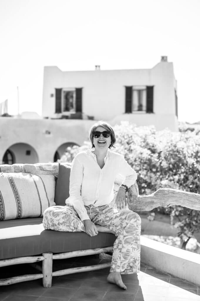 Vacances en Grèce : femme sur une terrasse cycladique Femme souriante en lin blanc sur une terrasse face à une villa cycladique en noir et blanc.